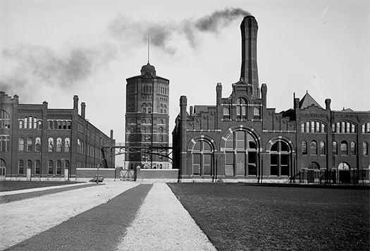 The front of a factory complex showing two workshops. Smoke rises from the chimney of one of the workshops, which sits astride an entry gate. A tall water tower sits further back, behind both workshops and the entry gate.