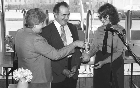Figure 15 As her husband looks on, a woman hands a ring of keys to a woman in park ranger uniform. All three are smiling. Behind them is a laundry counter and a plate-glass window overlooking a downtown street scene.
